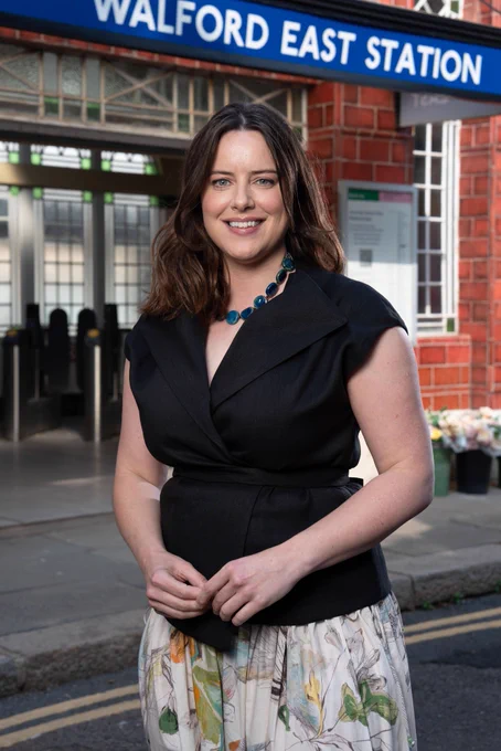A woman in a black top and floral skirt stands outside the fictional Walford East tube station smiling