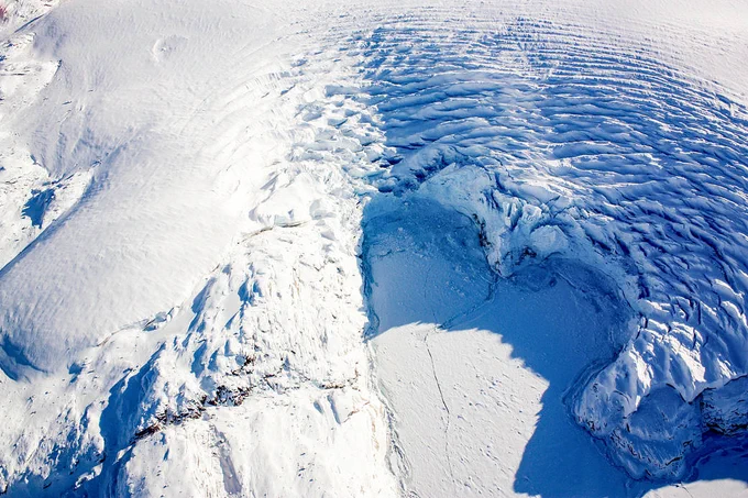 A photo of the calving front of a glacier in Greenland, as seen from an airplane in 2017. The ice has broken off in a way to make the front heart-shaped, with ridges rippling in the ice behind it.