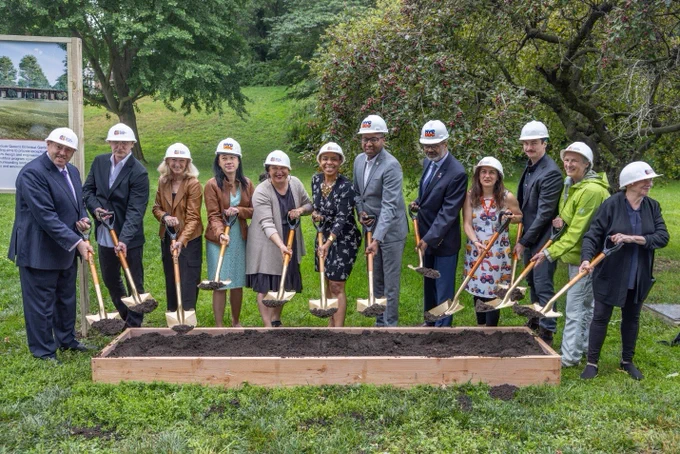 A photo of the groundbreaking ceremony, with a number of officials wearing hard hats and holding shovels over a bed of dirt. 