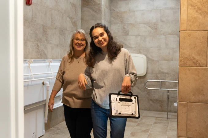 Mother and daughter smiling in a restroom with an adult-sized changing table in the background.