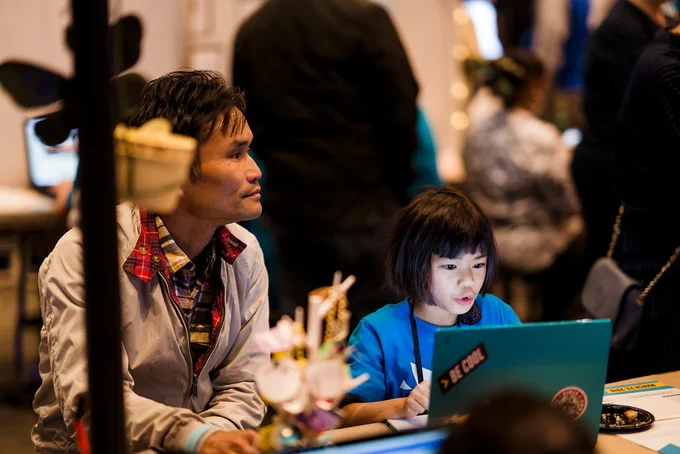 A young creator looking at a laptop with a mentor sat next to them. The laptop has a sticker on it that says 'Be Cool'.