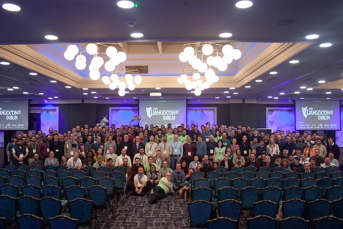 Group photo of DjangoCon Europe 2025 Dublin attendees, all looking at the camera from inside a ballroom, with chairs in the foreground