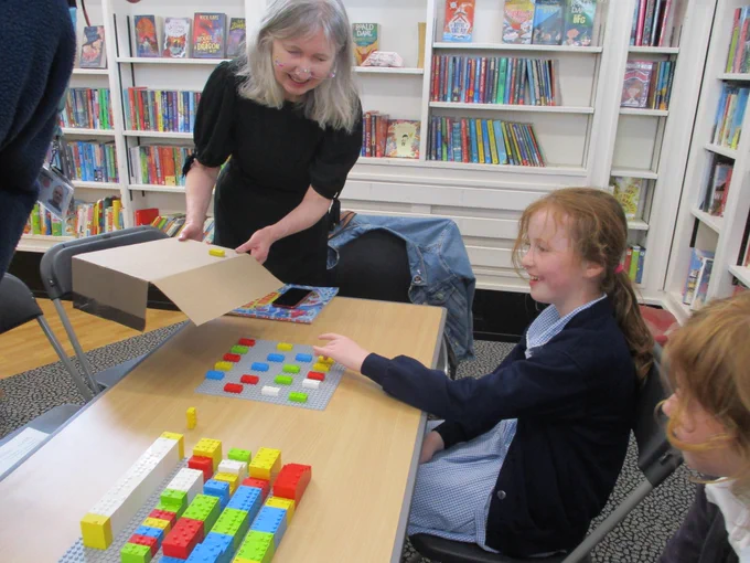 A girl is playing a game with the Braille Lego bricks and smiling. Her Grandma is watching and smiling over her.
