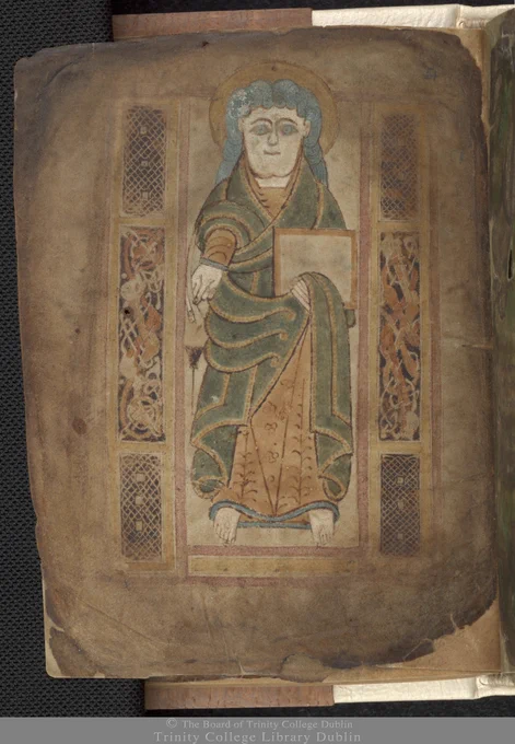 Portrait of St Matthew.  A decorated portrait page depicting a man - St Matthew - in a robe of green and gold, with blue hair and a halo.  He holds a book in his left hand within a fold in the robe.  He is surrounded by a frame containing interlacing and zoomorphic patterns