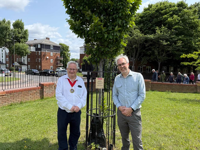 
The Mayor of Barnet and Grange Big Local Chair James, standing in front of a young tree in Oak Lane Open Space, East Finchley. This tree was planted in 2015 to mark the start of Grange Big Local. Both are white men with white hair, dress in smart shirts and trousers. 