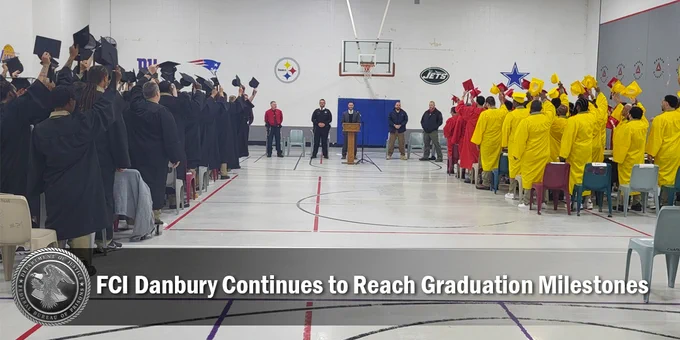 Graduation ceremony at FCI Danbury with attendees in caps and gowns, watching speakers at the front of a gymnasium.