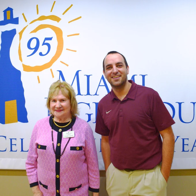 A photo of Virginia Jacko, President and Chief Executive Officer of Miami Lighthouse for the Blind & Visually Impaired, smiling for the camera with Dr. Michael Tuttle in front of the Miami Lighthouse for the Blind logo.