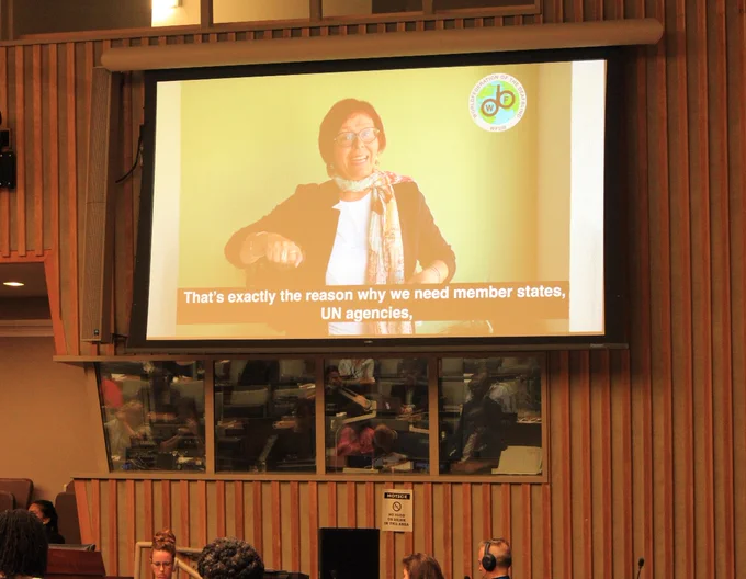 A large screen in a conference room displays a woman speaking in a video. She wears glasses, a scarf, and a black jacket, and is gesturing with her right hand. The video has subtitles that read: "That's exactly the reason why we need member states, UN agencies," and shows the logo of the World Federation of the Deafblind in the top-right corner. Below the screen, people are seated, some using headsets,