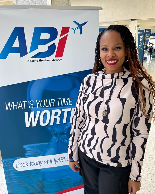 Person smiling and standing in front of a promotional banner for Abilene Regional Airport. The banner reads, "What's Your Time Worth? Book today at iFlyABI.com." The person is wearing a black and white striped top.