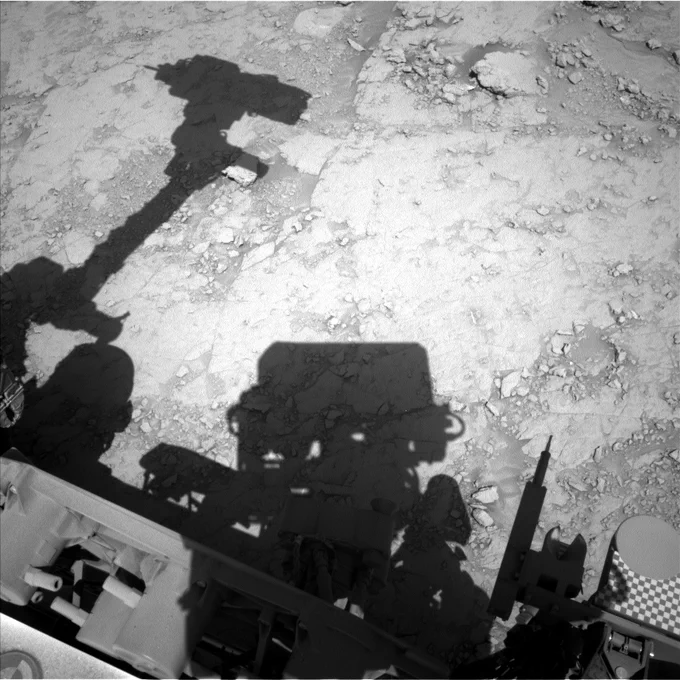 A black-and-white image shows the Curiosity rovers shadow on the surface of Mars. The rover has its robotic arm and drill extended out and the outline of the arm and tools is clear along the rocky surface.