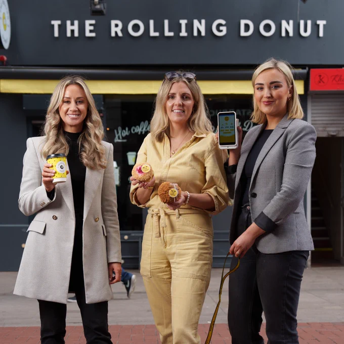 3 women stand outside the rolling donut shop holding coffee, donuts and a pay machine