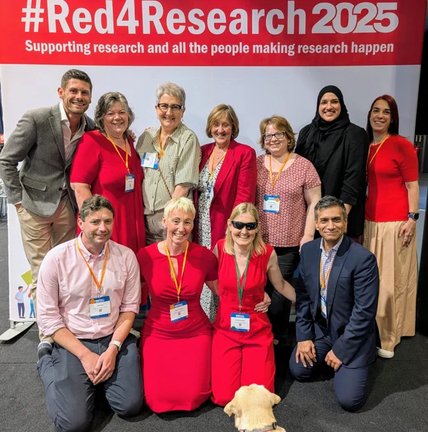A group of the R&D Forum Trustees and Working Group Chairs and Jessie the dog. Most of the group are wearing red and smiling at the camera in front of a #Red4Research banner.