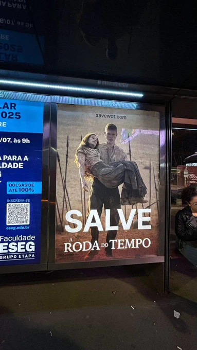 The image shows a billboard advertising the Brazilian film "Salve a Roda do Tempo" ("Save the Wheel of Time"). The billboard features a poster with a couple embracing, seemingly in a dramatic scene. The text on the billboard includes the film's title in large, bold lettering ("SALVE A RODA DO TEMPO"), the tagline "A RODA DO TEMPO" which translates to "The Wheel of Time", and website address "savewot.com". The billboard is located outdoors, likely near a bus stop or in a public area, in a city with streetlights illuminating it.  The background is dark, and other signs and streetlights are visible, indicating a nighttime setting.