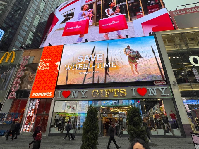 A vibrant cityscape with a large digital billboard displaying a person enjoying a vacation, with the slogan "Save the wheel of time" and a website address. Below the billboard, a storefront with the sign "I NYC GIFTS I NYC" is visible, flanked by smaller signs for Popeyes and McDonald's. People are strolling in front of the store, and tall evergreen trees adorn the sidewalk.