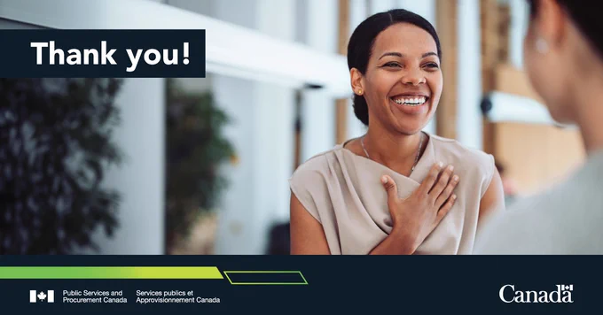 A person  stands with one hand on their chest, appearing to make a gesture of gratitude. The setting is indoors, with another person partially visible in the background. Text on the image: Thank you!