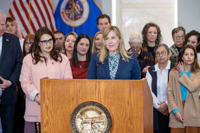 Speaker Emerita Melissa Hortman speaking at a podium with the Minnesota state seal at the PRO Act signing event, accompanied by a group of attentive listeners in the background.