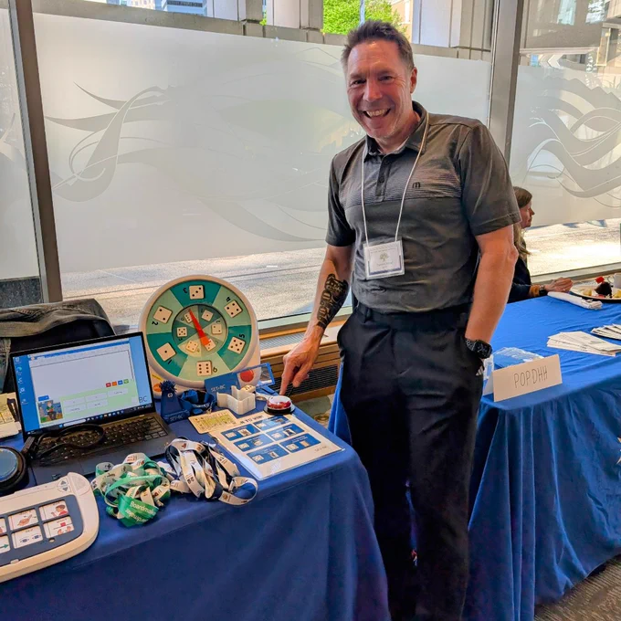 A man stands at a table pressing a switch beside an alternative spinner that are on a table with other assistive technologies.