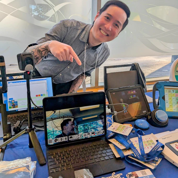 A man leans over a table and points to a laptop screen or the table top which has mini COREboards, switches, tablets and other assistive technologies.