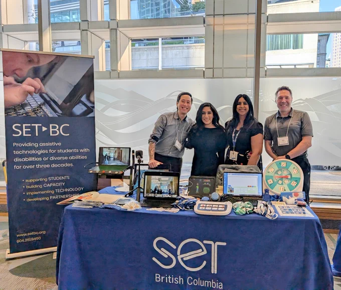 Four people stand behind a table with a blue tablecloth that reads SET British Columbia. Atop of the table are various type of assistive technology and resources. To the left is a SET-BC banner that reads "SET-BC Providing assistive technologies for students with disabilities or diverse abilities for over three decades."