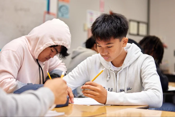 A group of students seated at a table together, each writing in a notebook.