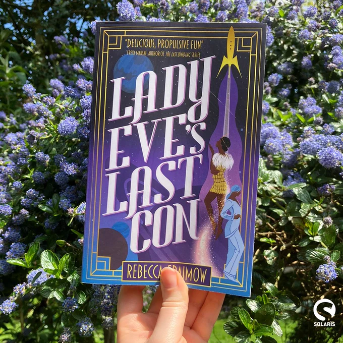 Against a background of blue California Lilac, a paperback copy of LADY EVE'S LAST CON by Rebecca Fraimow. The white Solaris logo is in the bottom right corner.