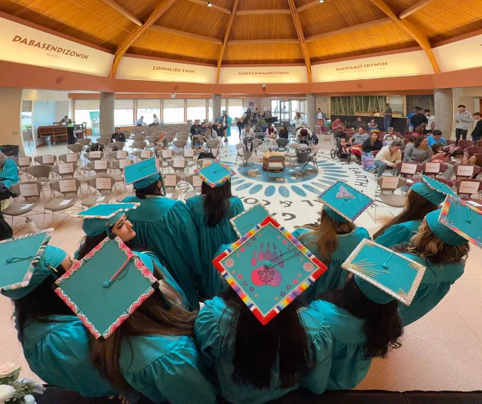 A photo of decorated graduation caps from the back as all of the graduates wearing them sit and face forward.