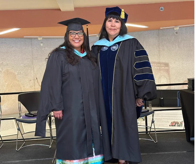 Two women stand in dark graduation robes, one wearing a doctoral tam.