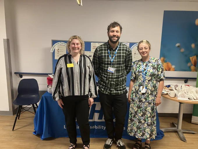 Staff members stand smiling in front of a blue table with “NHS Learning Disability Services” written on the cloth. Behind them is a display board with information on occupational therapy, psychology, and community nursing.