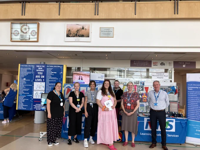 Staff members pose in front of an NHS Learning Disability Services stand inside Worcestershire Royal Hospital. There are colourful posters, a stall with bunting, and hospital signage in the background.