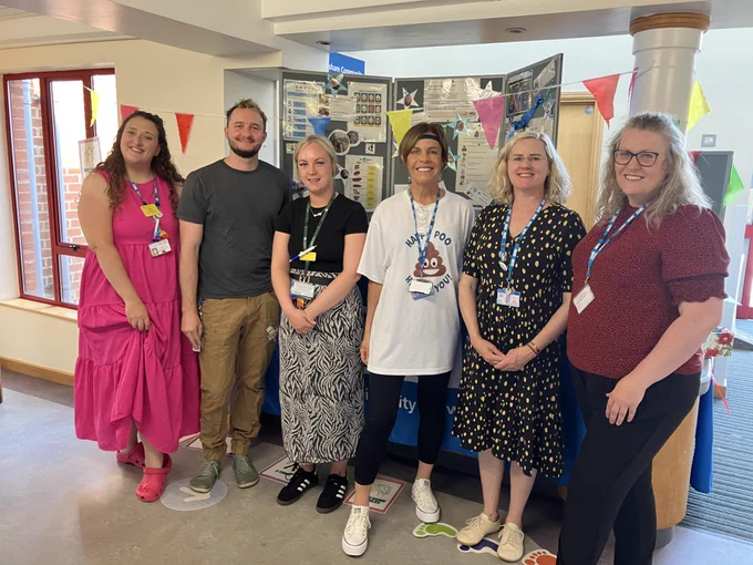 Staff members smile together at the Evesham Community Hospital event. The background is decorated with colourful bunting and informative health boards.