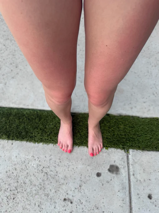 A close-up photo of Rowan McClane’s bare feet standing on a strip of artificial grass, with concrete on either side. Her toenails are painted bright pink. The image is taken from her top-down perspective, showing her legs up to the knees. The caption reads “Ready for summer!” with summer-related emojis and hashtags.