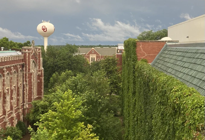Photo of OU campus from a 5th floor window, with views of several buildings, a water tower, and lots of greenery (tops of trees and vines on wall of library building).