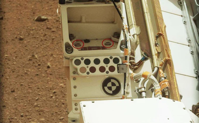 A close-up view of the Curiosity rover on Mars shows a section of the rover’s body that contains its calibration targets and two microchips (circled in red) mounted above them. These microchips contain the names of more than 1.2 million people who signed up to send their names to Mars. The dusty Martian surface is visible in the background.
