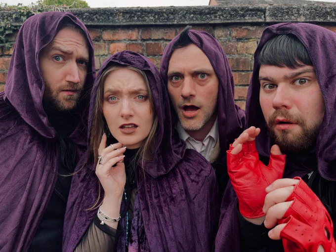 4 people looking suspiciously shocked down a camera wearing purple velvet cloaks with hoods. They stand in front of a brick wall. There are 3 men and one woman, and the man on the far right wears red fingerless gloves