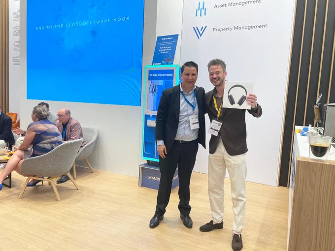 Two individuals smiling and posing at a conference booth with a background featuring banners about cloud software for property management. One person is holding an award.
