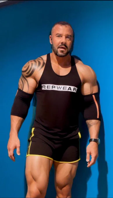 A heavily muscled man poses against a vivid blue background, wearing a tight black tank top labeled “RepWear” and short, body-hugging black shorts with yellow trim. His thick, muscular legs and arms are on full display, enhanced by tribal tattoos and compression sleeves, exuding raw power and sexual energy.