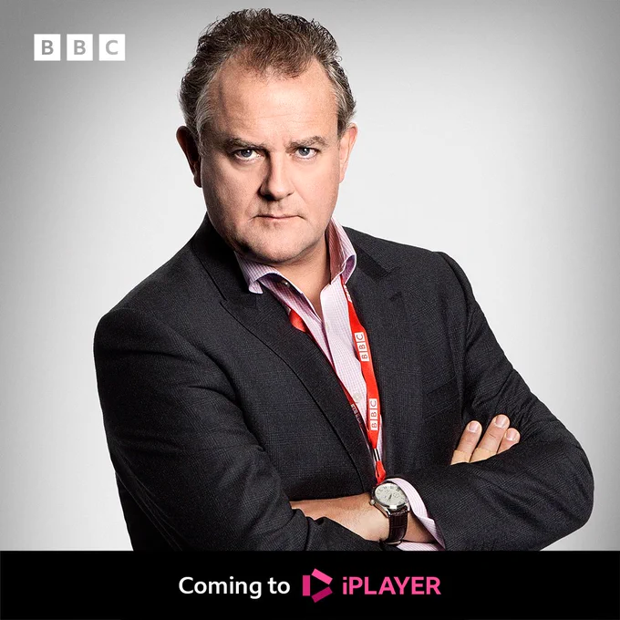 A man in a suit stands with his arms crossed looking serious