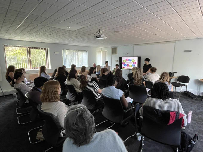 Image shows a room full of people sitting in chairs looking at a screen