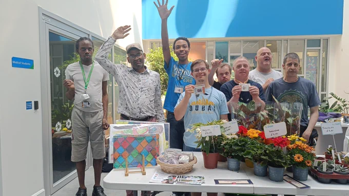 A group of smiling Share students posing behind their market stall. There is a table  covered with vibrant plants in green, white, yellow, orange, and red, plus Share art prints, greeting cards, and leaflets.