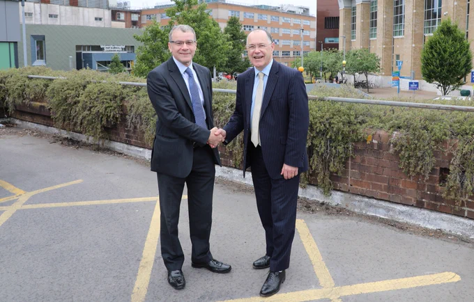 Chief executive at University Hospital Southampton, David French, and University of Southampton Vice-Chancellor shake hands. Behind them is a hospital building with a sign that reads "University Hospital Southampton." Greenery and trees are visible in the background.