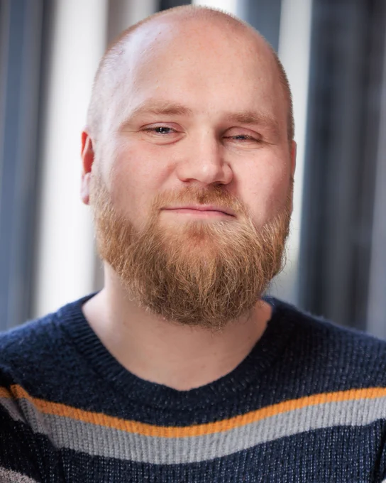 A headshot of a man with a ginger beard and shaved head. He smiles slightly and is wearing a striped jumper.