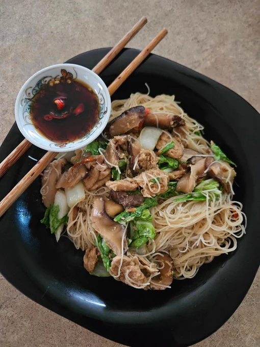 Dinner as described served on a squareish black soup plate. A pair of wooden chopsticks lie atop the plate on a diagonal, and a small dish with sliced red chilli & soy sauce is balanced on the chopsticks.