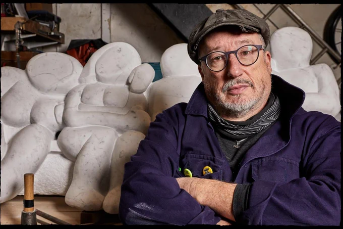 Tony HEaton, a white man in blue overalls stands cross armed in front of a marble sculpture