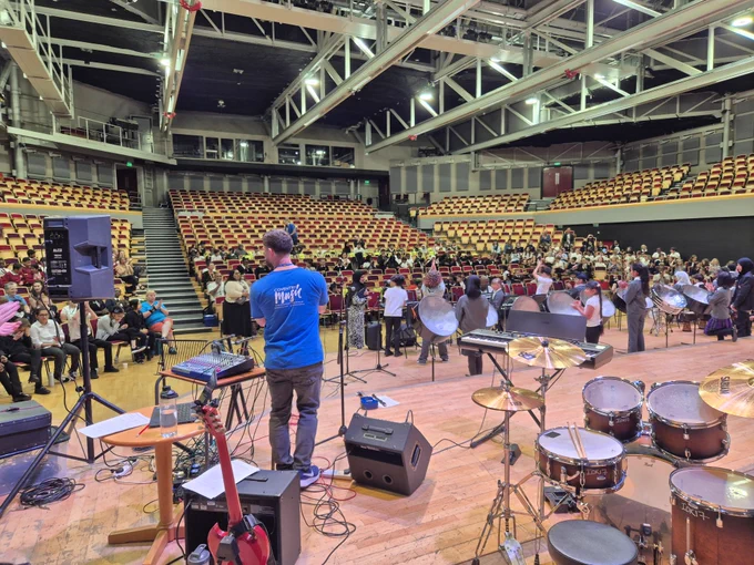 Music equipment on stage in an auditorium. There are people seated in the theatre-style seats and a member of Coventry Music staff is stood on the stage.