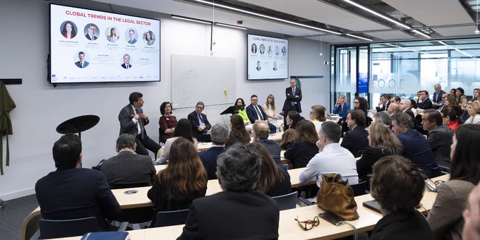 Panel discussion at a conference on global trends in the legal sector, with seven people seated and standing in front of a screen displaying their headshots and an audience listening attentively.
