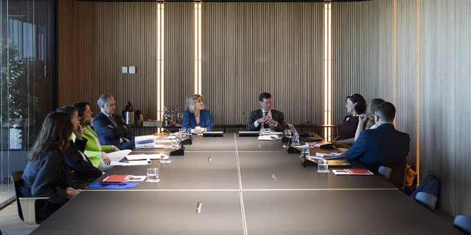 A group of professionals sitting around a long conference table in a well-lit room with wooden walls and modern design, engaged in a meeting.