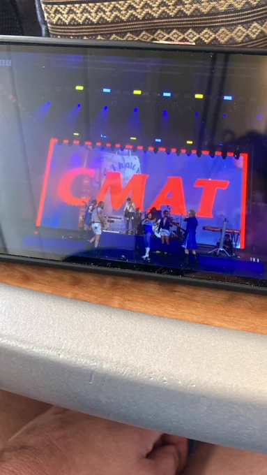 Photo of my phone screen on the train table. On the screen is CMAT playing Glastonbury with the letters C M A T in big red letters