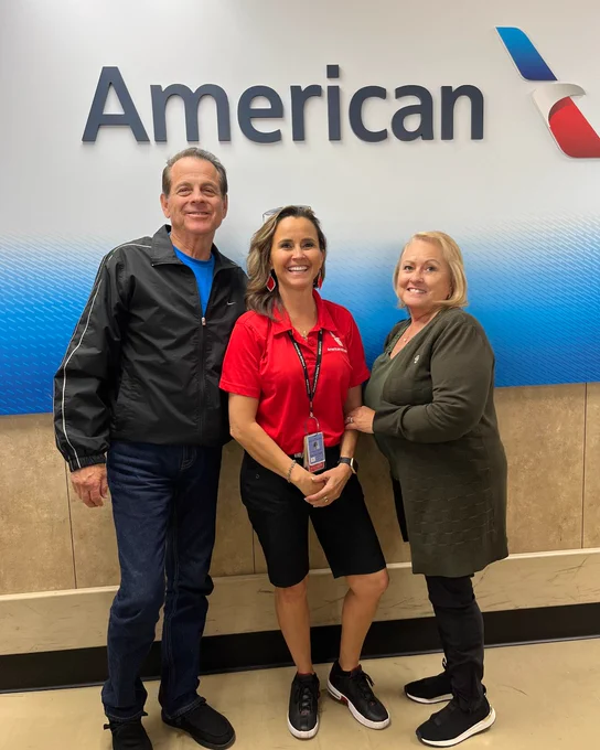 Three individuals standing and smiling in front of a wall with the American Airlines logo.