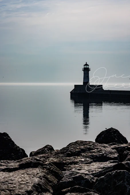Lighthouse on the water with rocks in the foreground. Light blue foggy sky.