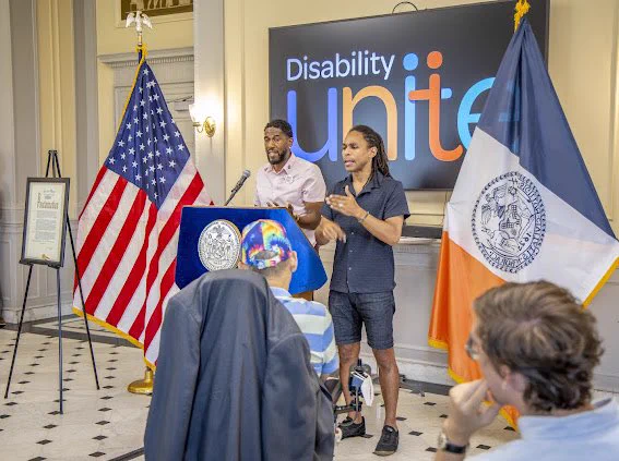 The Public Advocate speaks from behind a blue lectern while a translator uses ASL next to him